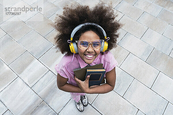 Lächelndes Schulmädchen mit Kopfhörern  das ein Buch hält und auf dem Fußweg steht