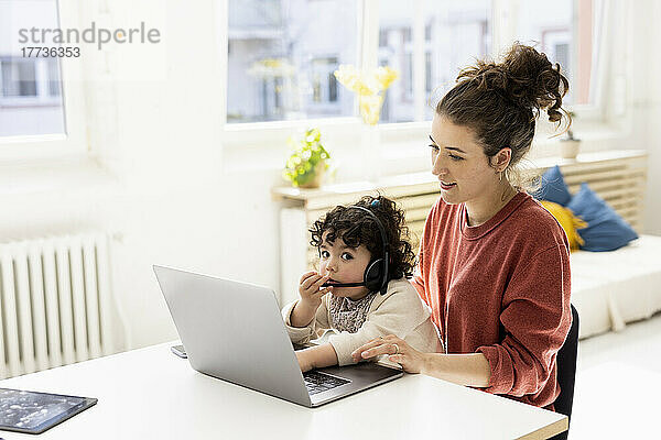 Kleines Mädchen mit Headset sitzt auf dem Schoß der Mutter und benutzt Laptop