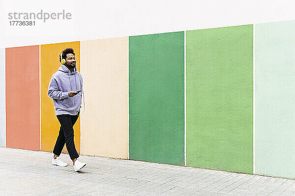 Mann hält Mobiltelefon in der Hand und hört Musik über Kopfhörer  während er auf Fußweg an einer bunten Wand entlang läuft