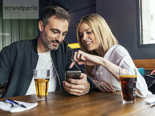 Lächelnde Frau zeigt auf das Smartphone  das ein Mann am Tisch im Restaurant hält