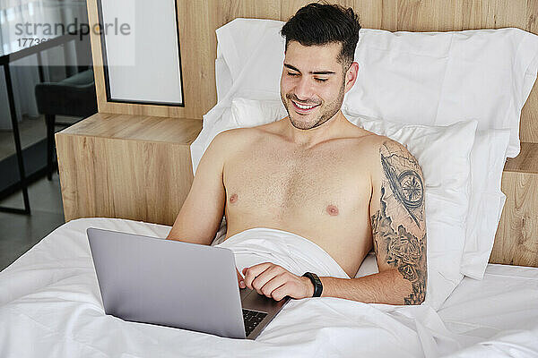 Lächelnder Mann ohne Hemd benutzt Laptop im Bett zu Hause