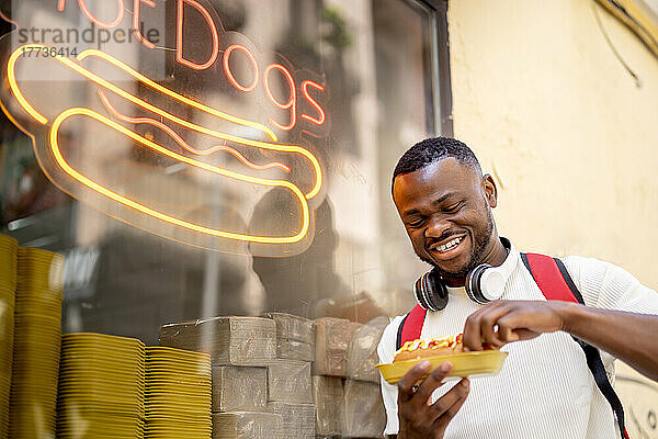 Glücklicher junger Mann  der Hot Dog isst und sich an das Fenster des Restaurants lehnt