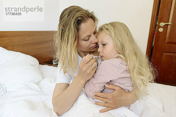 Mutter trägt Tochter und misst Temperatur mit Thermometer im Schlafzimmer