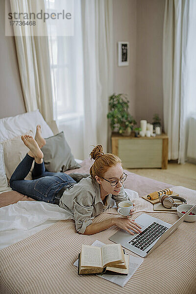 Lächelnde Frau mit Kaffeetasse und Laptop  die zu Hause im Bett liegt