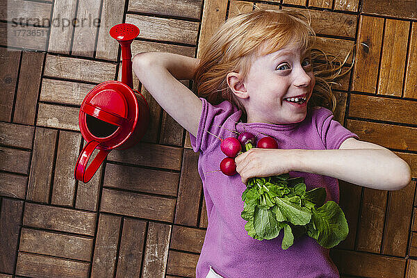 Happy girl with radish lying by watering can on balcony floor