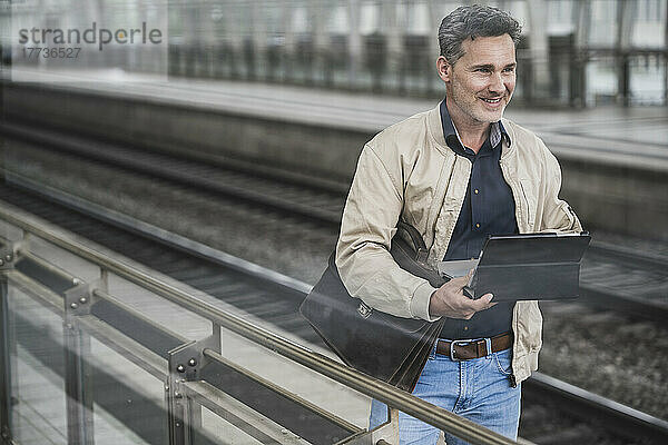 Lächelnder Geschäftsmann mit Tablet-PC und Tasche steht am Bahnhof