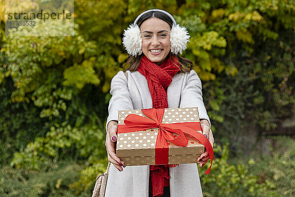 Lächelnde Frau mit Ohrenschützer überreicht Weihnachtsgeschenk im Park