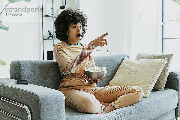 Schockierte Afro-Frau sitzt vor dem Fernseher auf dem Sofa und zeigt mit einer Schüssel Popcorn nach Hause