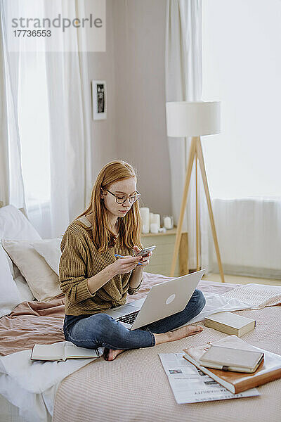 Frau schreibt Textnachrichten per Smartphone und sitzt mit gekreuzten Beinen und Laptop zu Hause im Bett