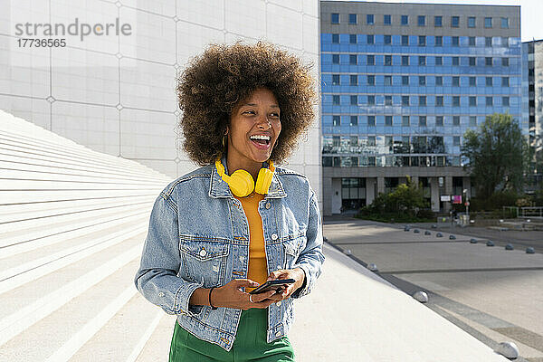 Fröhliche Afro-Frau mit kabellosen Kopfhörern steht an einem sonnigen Tag mit ihrem Smartphone neben Schritt