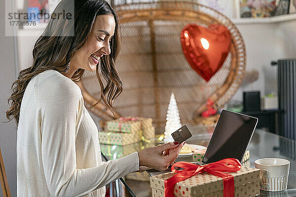 Glückliche junge Frau mit Kreditkarte sitzt mit Laptop und Weihnachtsgeschenk am Tisch