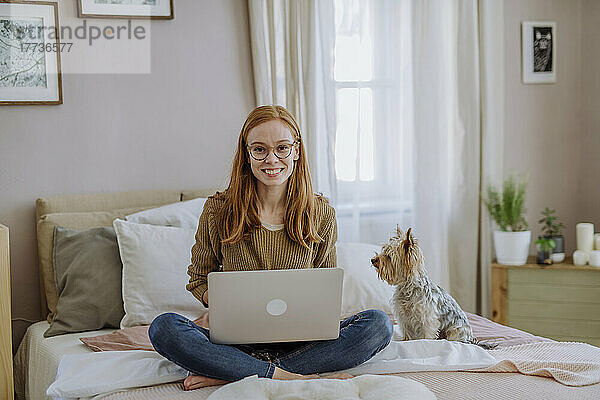 Lächelnde Frau sitzt mit Laptop neben ihrem Hund zu Hause im Bett