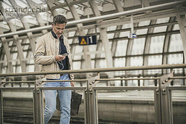 Reifer Geschäftsmann benutzt Smartphone und läuft mit Tasche am Bahnhof