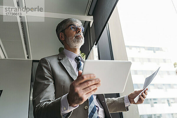 Reifer Geschäftsmann mit Dokumenten und Tablet-PC am Fenster im Büro