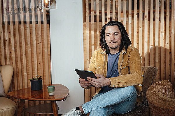 Lächelnder Hipster-Mann mit Tablet-PC sitzt auf einem Stuhl im Café
