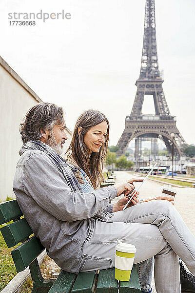 Lächelnde Frau und Mann mit Tablet-PC sitzen auf einer Bank in der Nähe des Eiffelturms  Paris  Frankreich