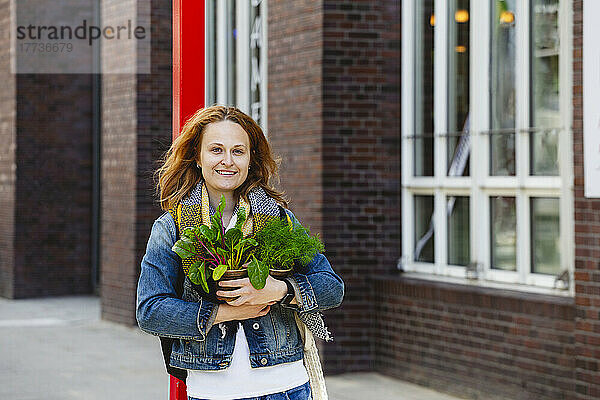 Lächelnde Frau steht mit Dill- und Mangoldpflanzen vor dem Gebäude