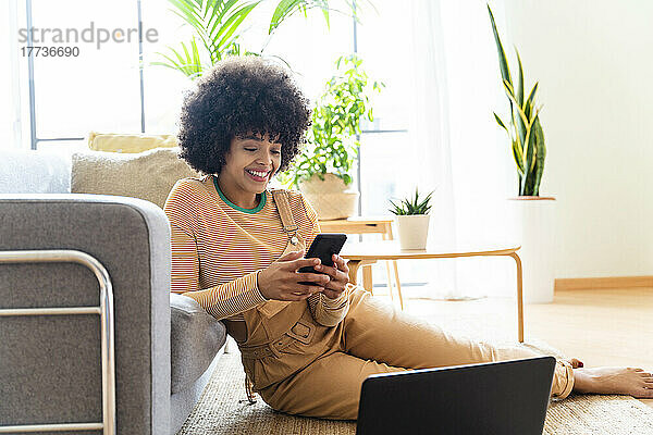 Smiling woman using smart phone sitting with laptop by sofa at home