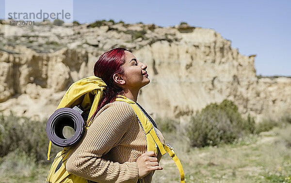 Lächelnde Frau mit Rucksack steht an einem sonnigen Tag mit geschlossenen Augen