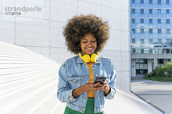 Lächelnde Frau mit Afro-Frisur  die auf der Treppe SMS über ihr Mobiltelefon sendet