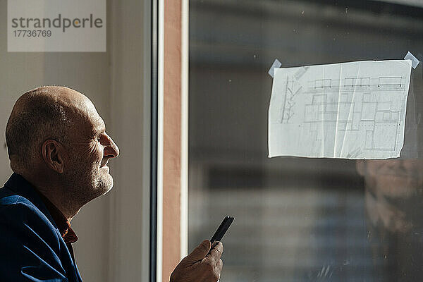 Geschäftsmann hält Mobiltelefon und analysiert Blaupause am Fenster im Büro