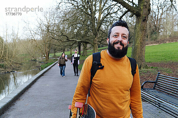 Lächelnder Mann mit Skateboard und Freunden  die im Hintergrund im Park spazieren gehen