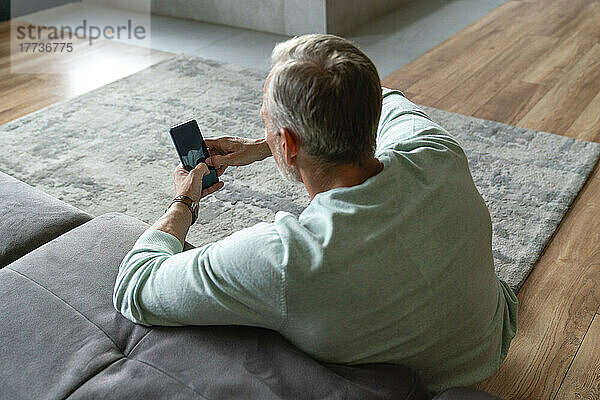 Mature man using smart phone leaning on sofa in living room at home