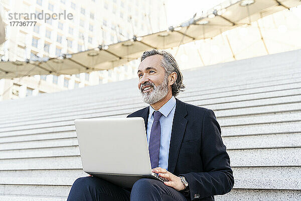 Glücklicher Geschäftsmann mit Laptop  der auf Stufen sitzt