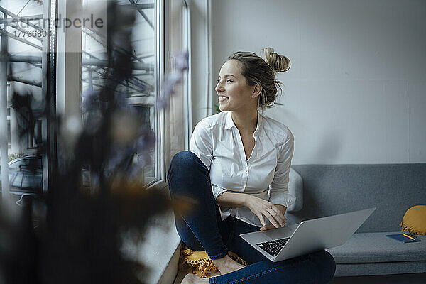 Lächelnde Geschäftsfrau mit Laptop sitzt auf der Witwenbank im Büro