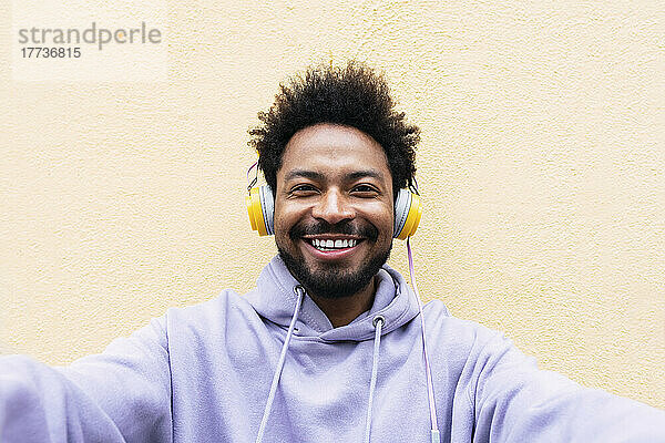 Lächelnder Mann  der über Kopfhörer Musik hört und ein Selfie vor einer cremefarbenen Wand macht