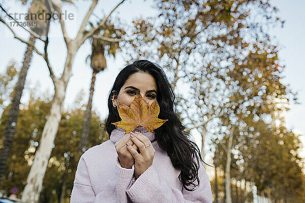 Junge Frau bedeckt Gesicht mit Herbstblatt im Park