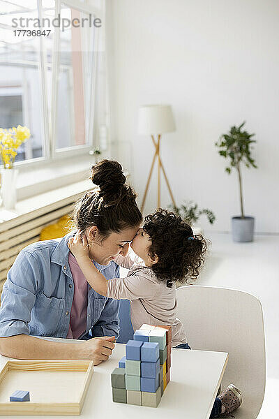 Tochter küsst Mutter auf die Stirn  die mit Bausteinen am Tisch sitzt