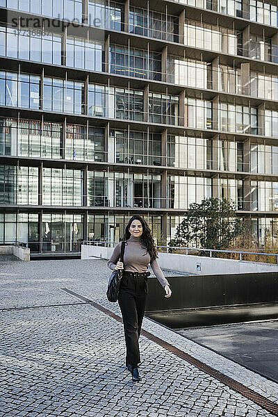 Lächelnde junge Geschäftsfrau  die vor einem modernen Gebäude im Finanzviertel läuft