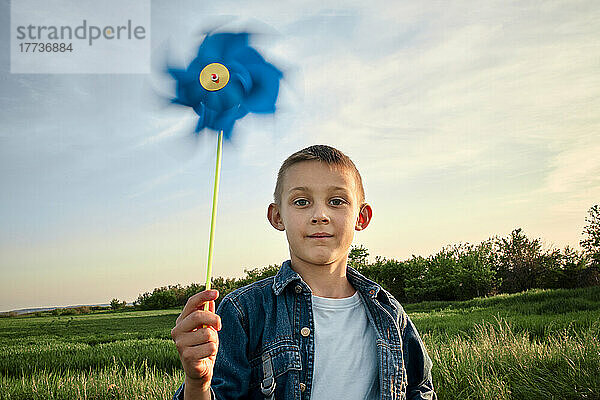 Netter Junge hält bei Sonnenuntergang ein blaues Windradspielzeug auf dem Feld