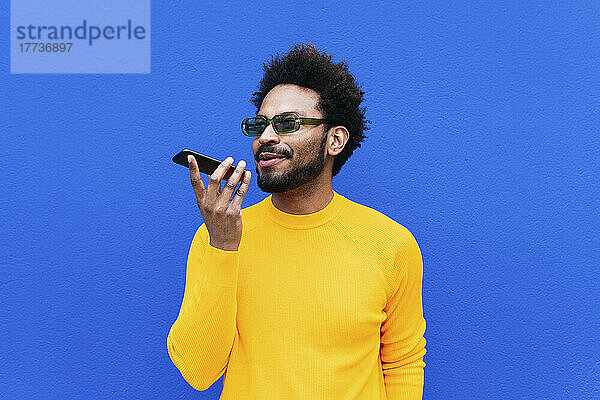 Lächelnder Mann  der über einen Lautsprecher vor einer blauen Wand mit seinem Mobiltelefon telefoniert