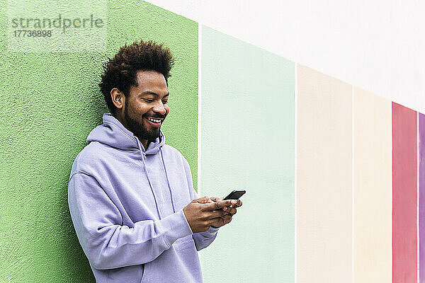 Glücklicher Mann benutzt Smartphone und lehnt an bunter Wand