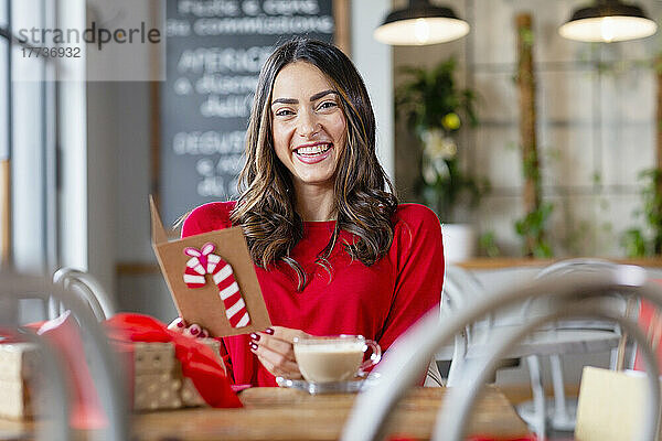 Fröhliche Frau mit Weihnachtskarte sitzt am Tisch im Café