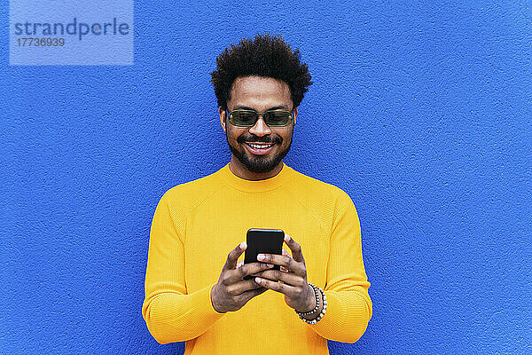 Lächelnder Afro-Mann benutzt Smartphone vor blauer Wand