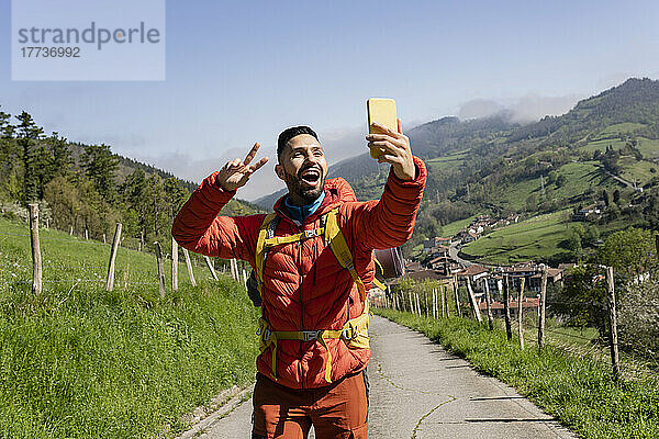 Glücklicher Mann gestikuliert Friedenszeichen und macht an einem sonnigen Tag ein Selfie mit dem Smartphone