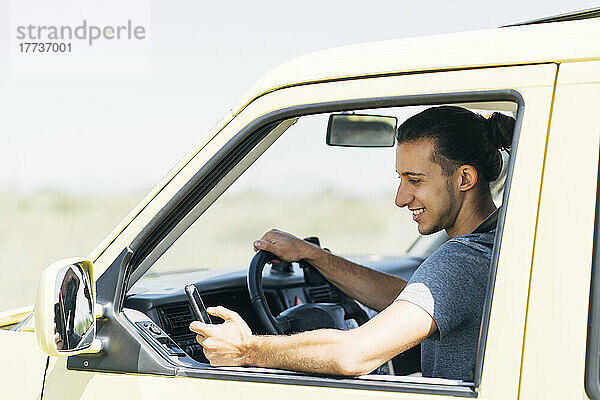 Lächelnder junger Mann  der an einem sonnigen Tag im Van sitzt und sein Smartphone benutzt