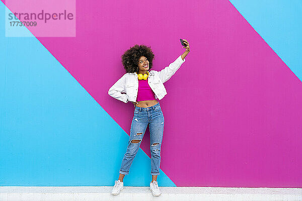 Lächelnde Frau mit der Hand auf der Hüfte macht ein Selfie mit dem Mobiltelefon vor einer rosa und blauen Wand