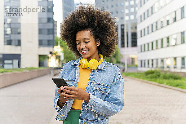 Lächelnde Afro-Frau  die Textnachrichten über ihr Smartphone sendet und auf dem Fußweg steht