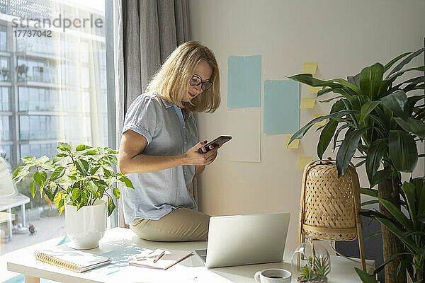 Blonde Geschäftsfrau mit Brille und Smartphone sitzt am Schreibtisch im Heimbüro