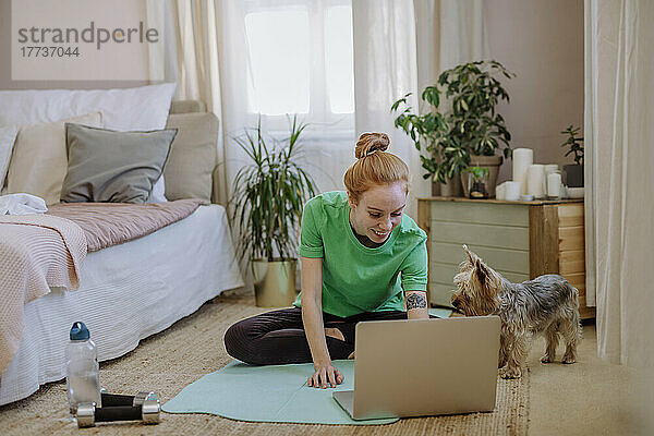 Lächelnde Frau schaut sich Online-Tutorial über Laptop an  während sie zu Hause neben ihrem Hund auf der Trainingsmatte sitzt