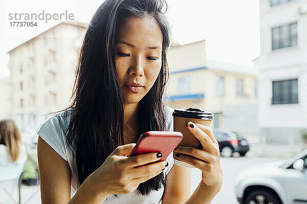 Schöne Frau hält eine wiederverwendbare Kaffeetasse in der Hand und sendet SMS über ihr Smartphone