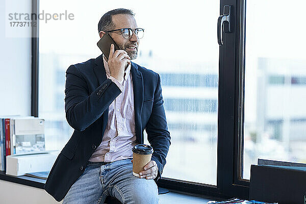 Geschäftsmann hält Einweg-Kaffeetasse in der Hand und spricht mit Smartphone am Fenster im Büro