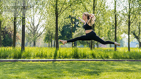 Junge Frau springt mit ausgestreckten Armen auf der Wiese im Park