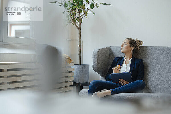 Nachdenkliche Geschäftsfrau mit Tablet-PC sitzt auf dem Sofa im Büro