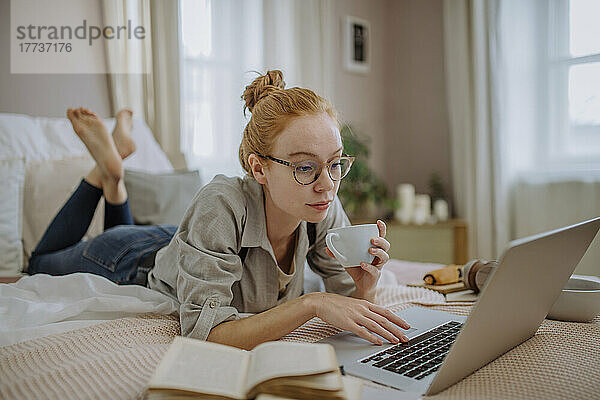 Frau mit Kaffeetasse und Laptop liegt zu Hause im Bett