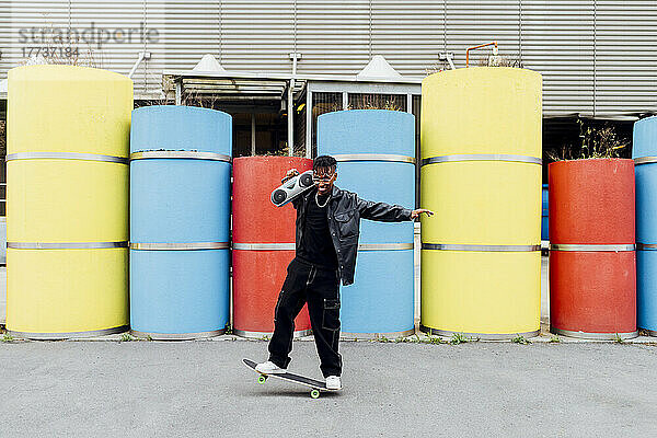 Glücklicher junger Mann mit Ghettoblaster balanciert auf Skateboard vor Betonrohren